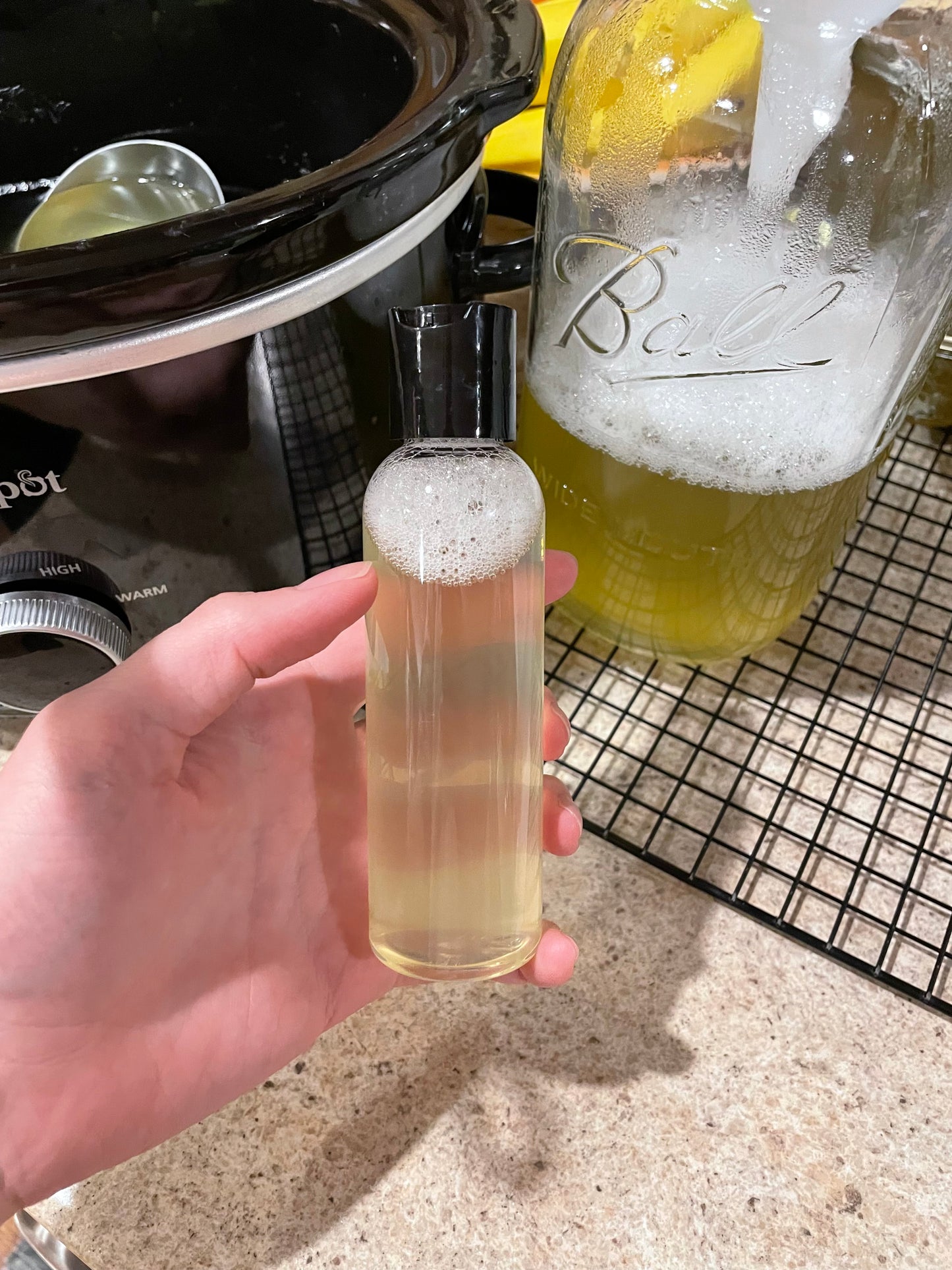 A hand holding a small snap top container with liquid soap inside. The liquid is a clear creamy color. The background is a kitchen countertop.