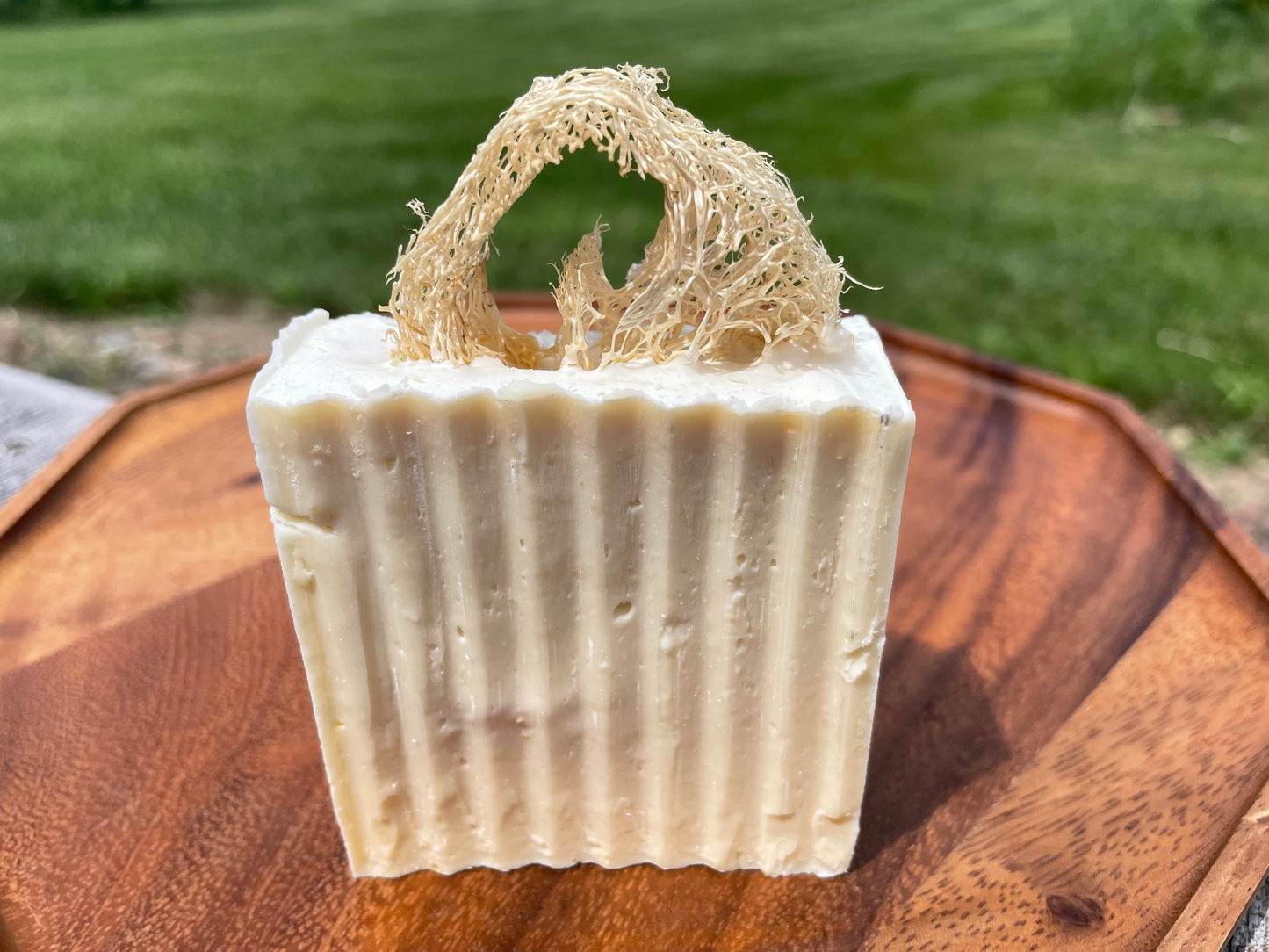 Bar of soap with a natural loofah on top, placed on a wooden surface with grass in the background.