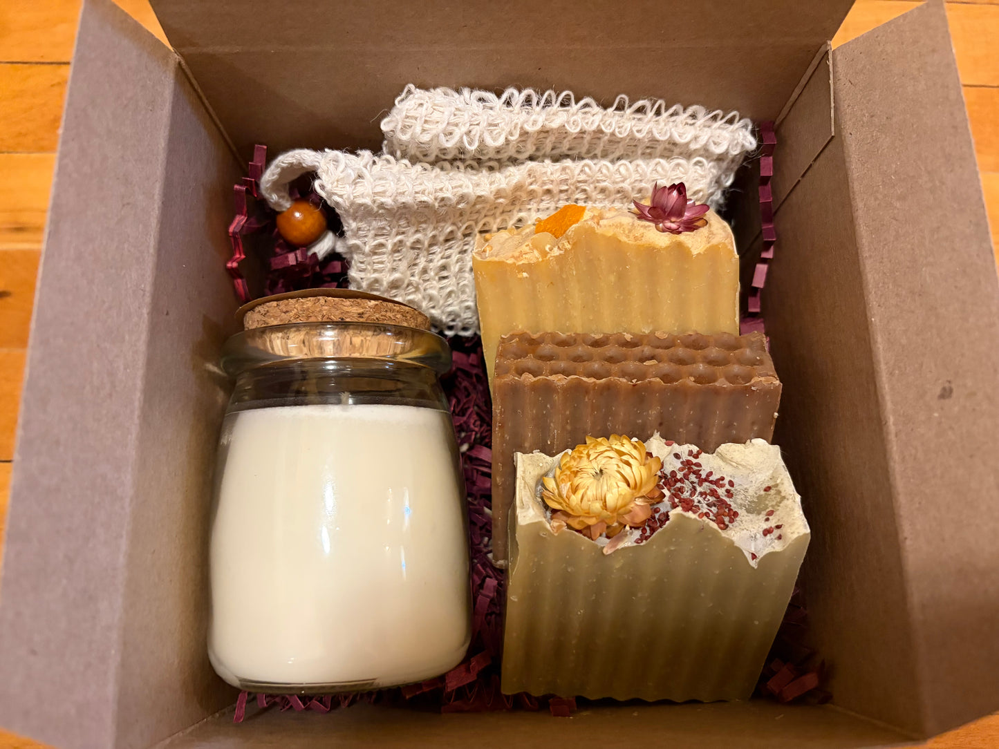 Gift set box with a candle jar, three bars of soap and a sisal soap loofa bag in a box on a wooden surface. Hard soap bars are a variety of colors including yellow, brown and green.