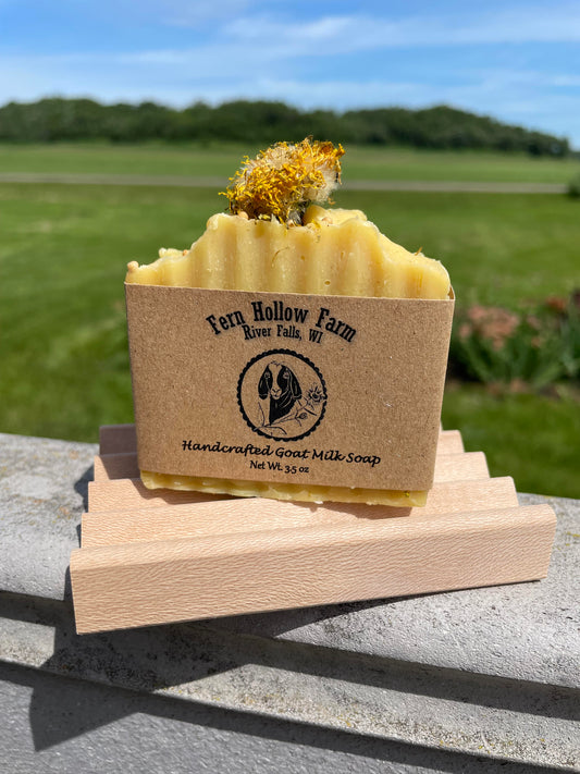 Rectangle bar of soap that is yellow in color with a dried dandelion on top. It is photographed outside and is located on a wood soap dish with a grassy background.