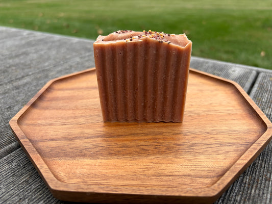 A bar of dark pink soap sitting on a wooden tray outside. The bar is topped with seeds. 