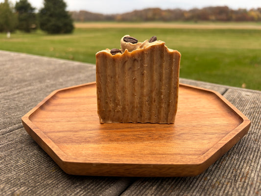 A bar of brown soap sitting on a wood tray outside. Coffee beans are on the top of the soap. 