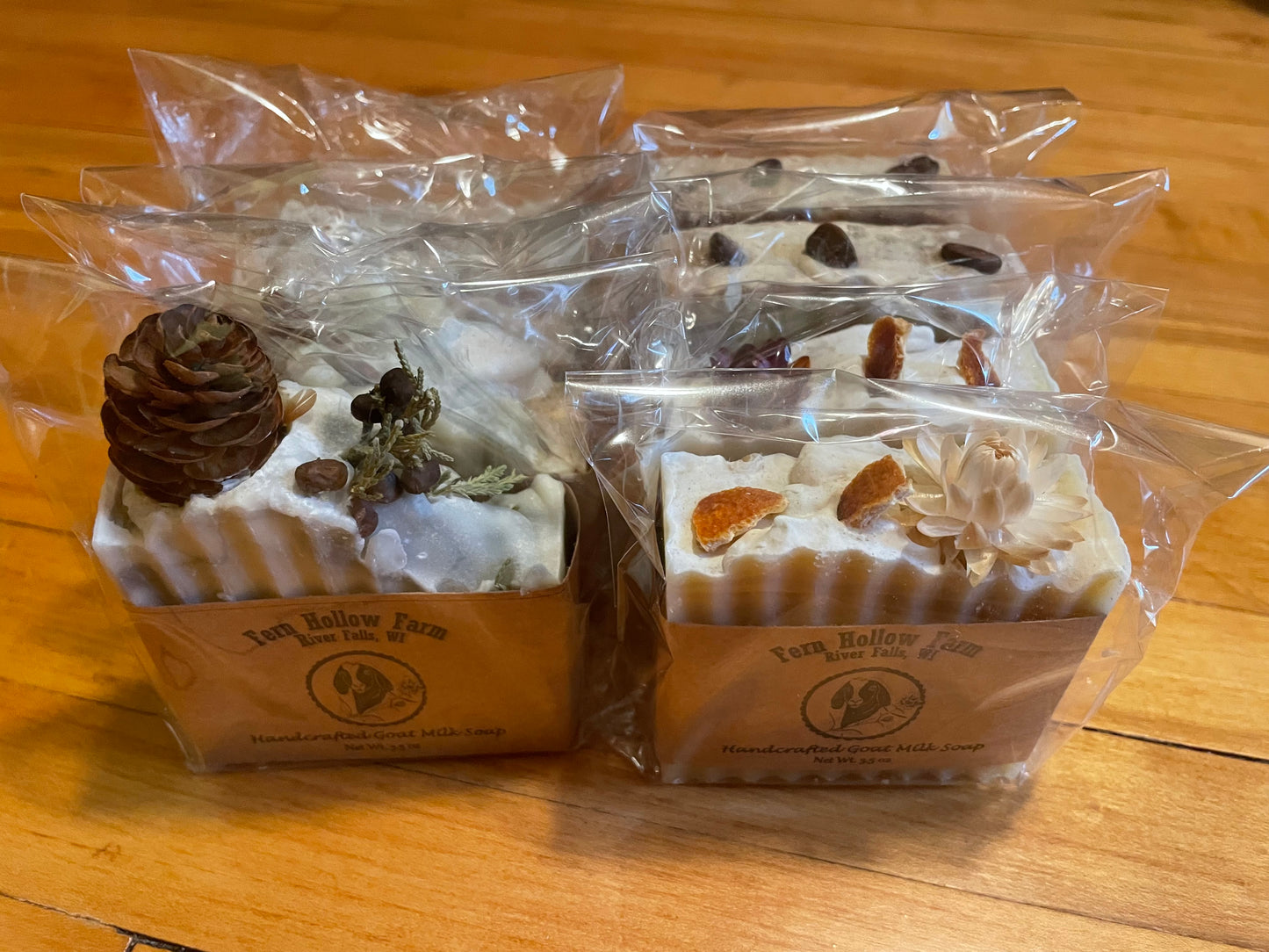 A collection of packaged bar soaps with natural decorations, such as herbs and pine cones, displayed on a wooden surface.