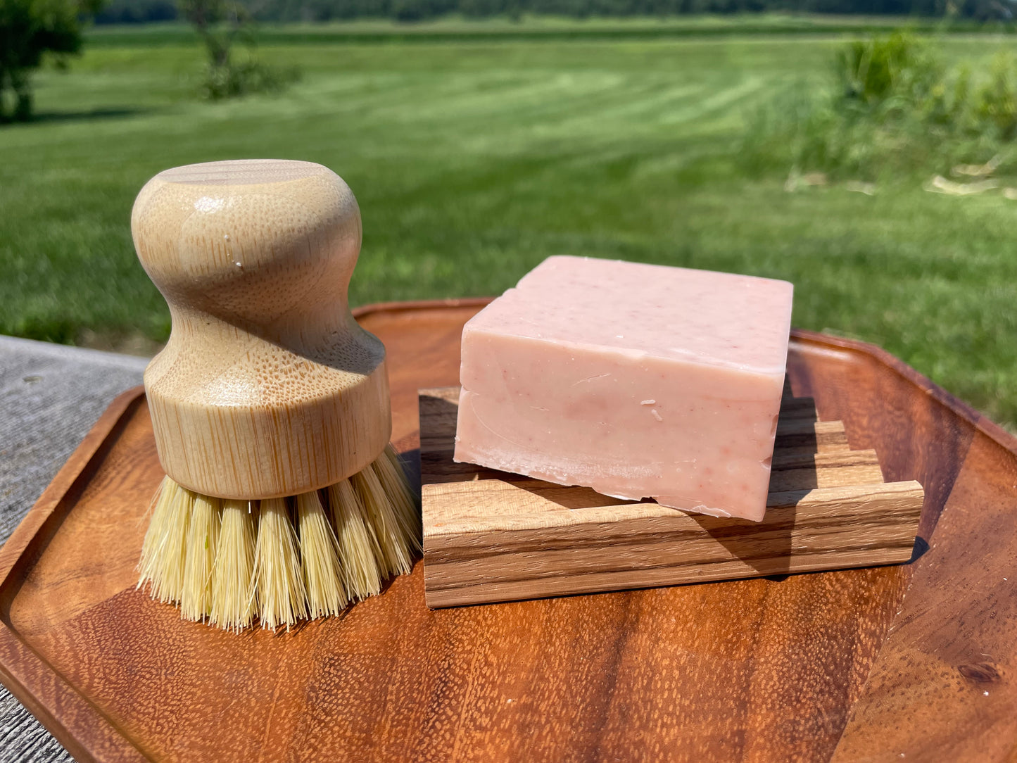 Wooden brush and pink colored soap on a wooden tray with a grassy field background