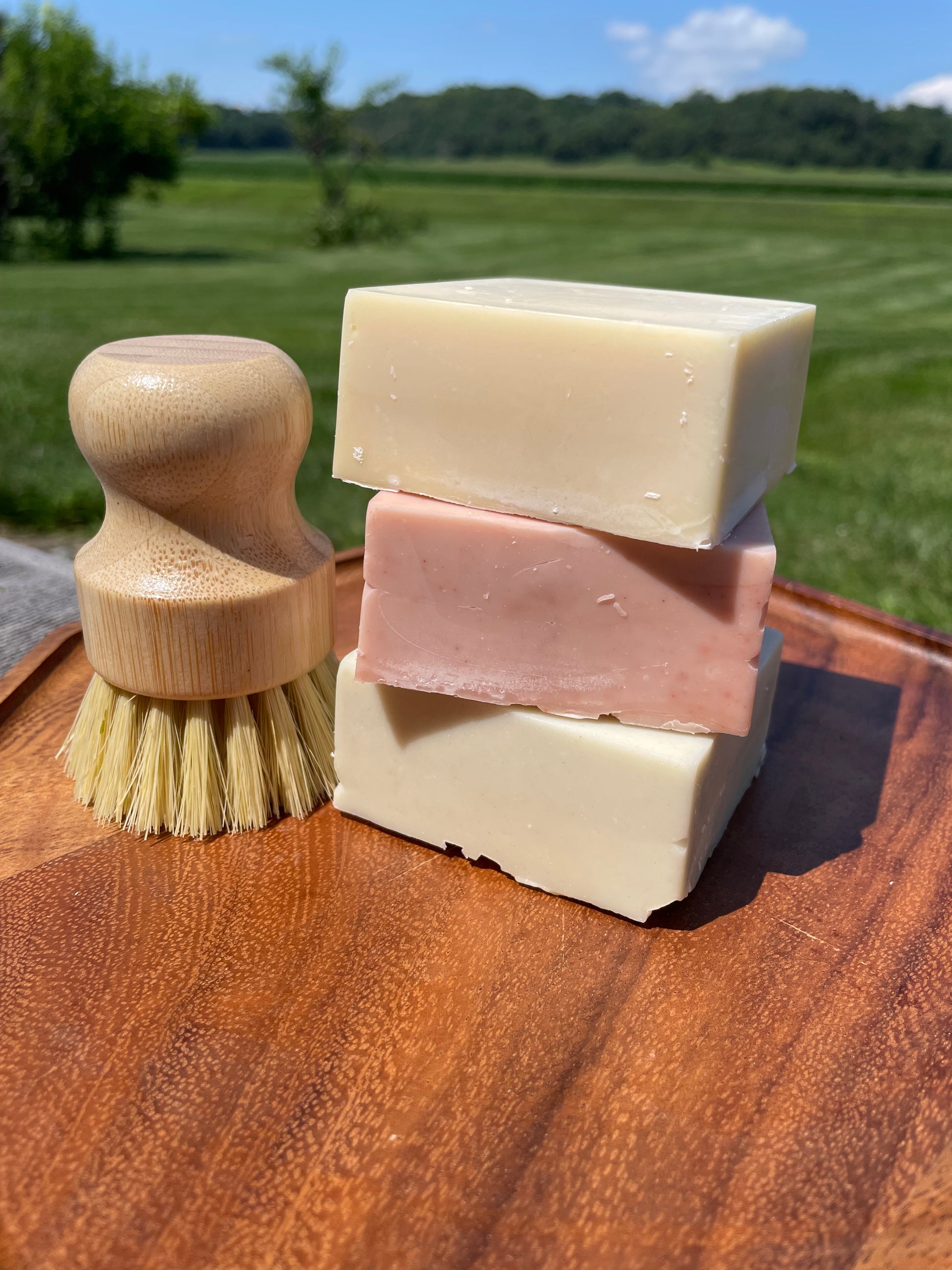 Three bars of soap and a brush on a wooden surface with a natural landscape in the background
