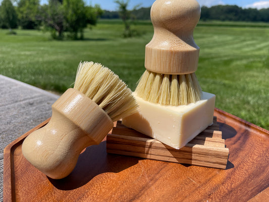Two wooden dish soap brushes with bamboo bristles on a wooden plate. Brushes are sitting on a square soap bar and wood soap dish.