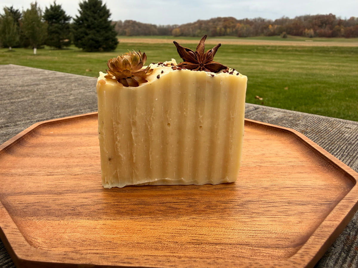 A bar of white soap on a wooden tray outside. The soap is topped with a strawflowers, star anise and seeds. 