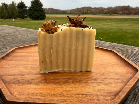 A bar of white soap on a wooden tray outside. The soap is topped with a strawflowers, star anise and seeds. 