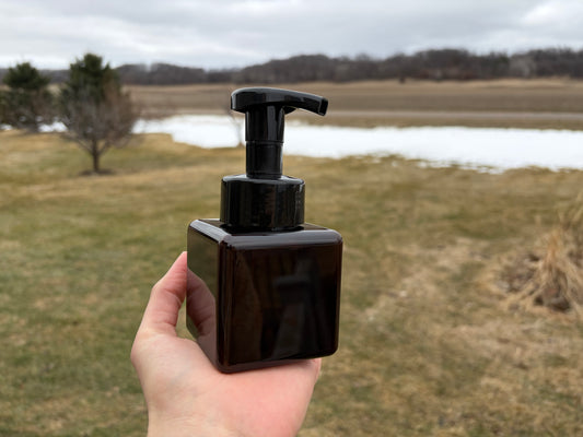 Amber brown foaming soap dispenser held in a hand outside with a grass yard and a small amount of snow in the background.