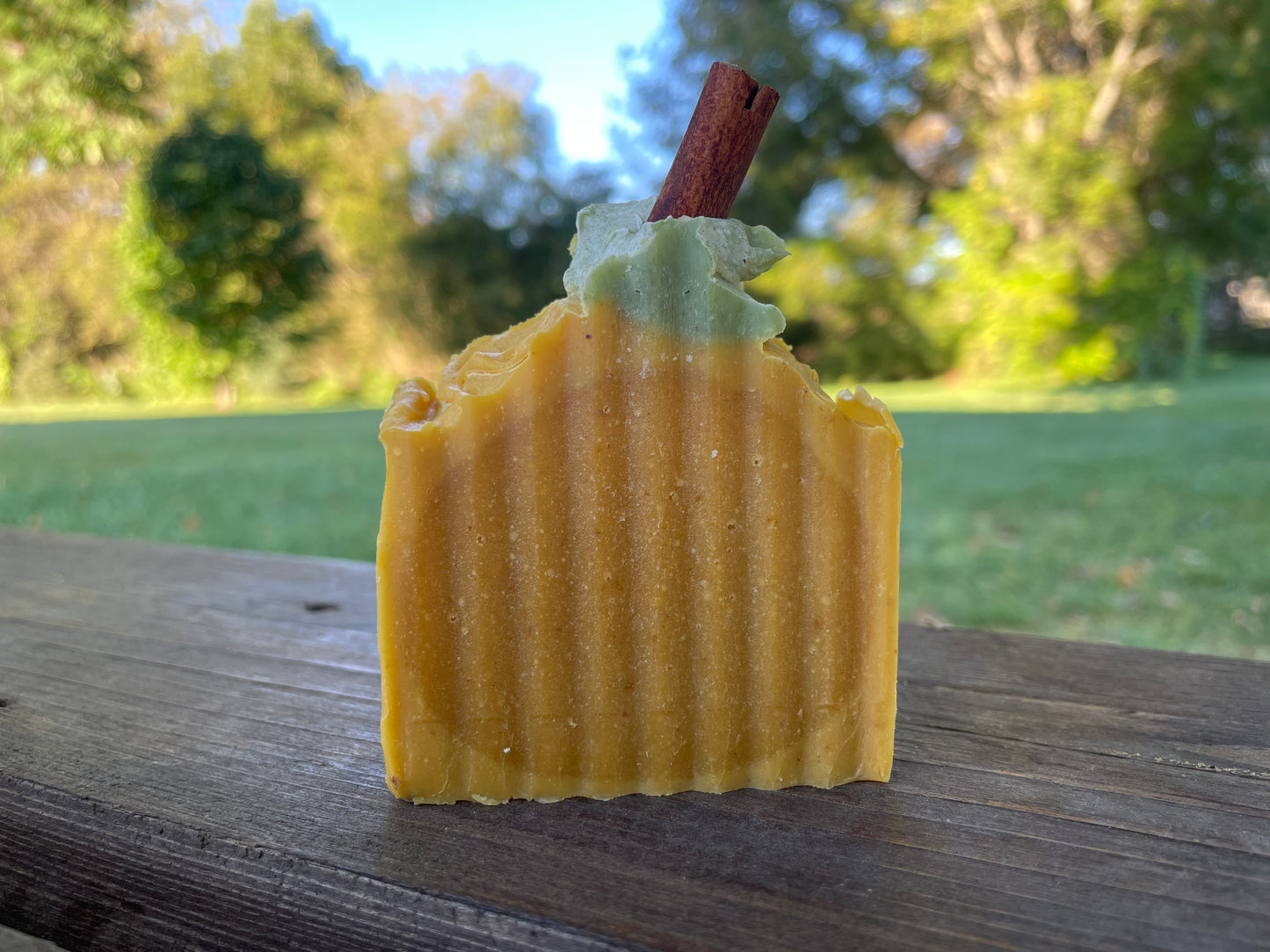 A bar of orange solid soap that is designed to look like a pumpkin. It is orange with a bit of green soap on top with a piece of cinnamon stick poking out the top. There is no label on the bar of soap. The photo is set in an outdoor setting with the soap on a wood table and grass in the background. 