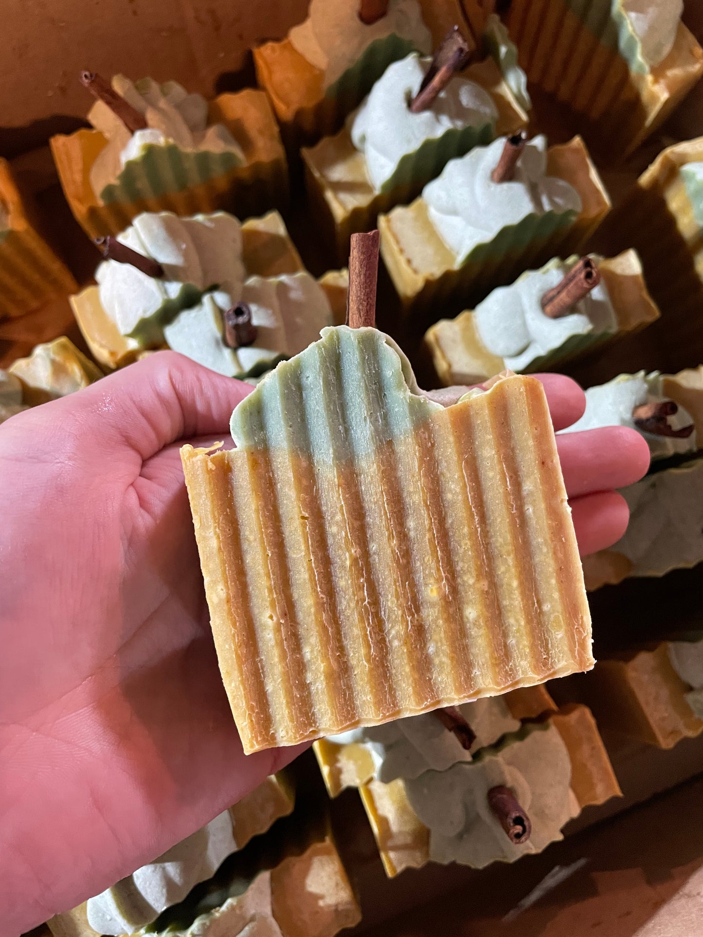A bar of orange solid soap that is designed to look like a pumpkin. It is orange with a bit of green soap on top with a piece of cinnamon stick poking out the top. There is no label on the bar of soap. The soap is held in a hand and matching bars are present in the background. 