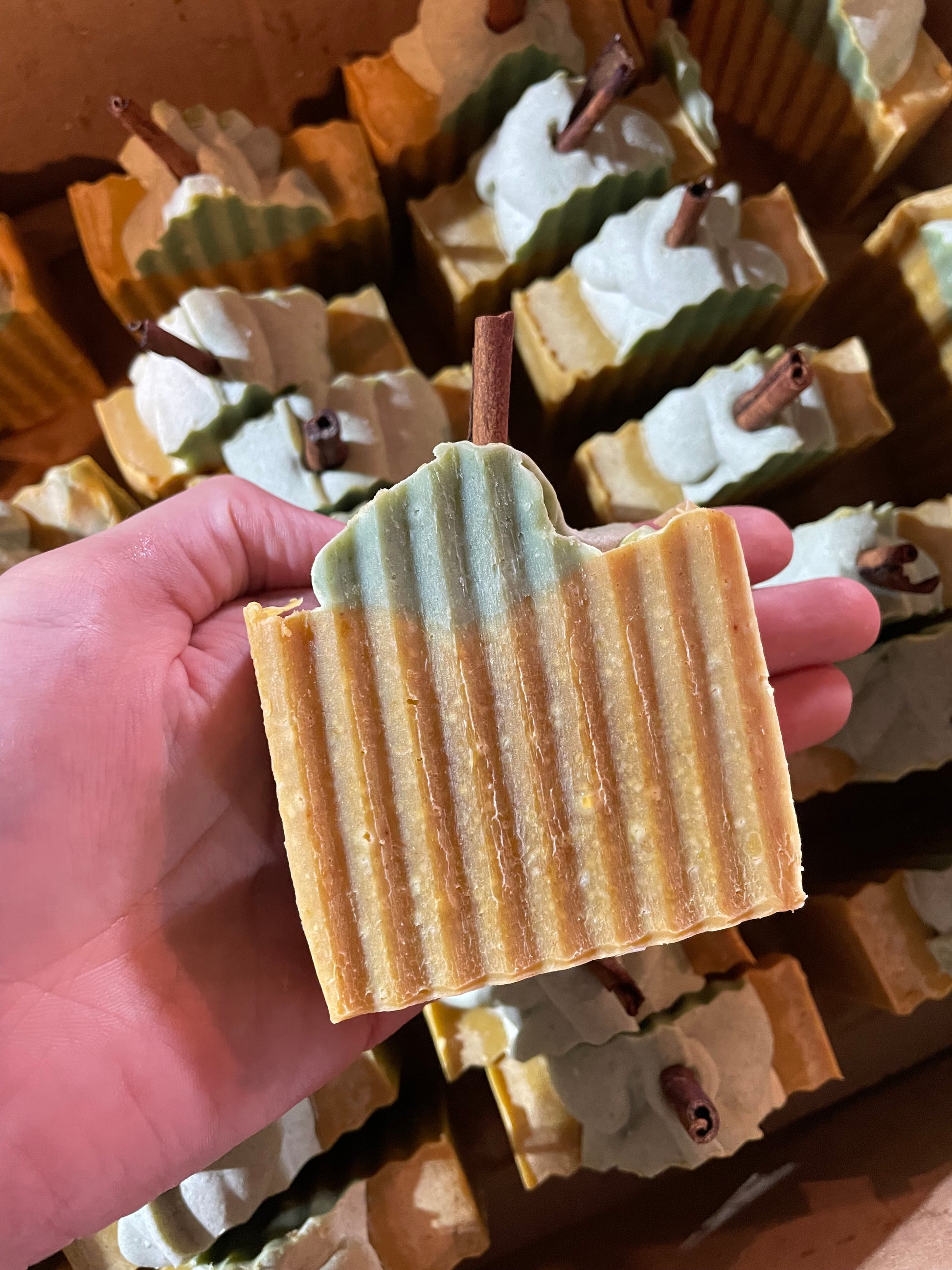 A bar of orange solid soap that is designed to look like a pumpkin. It is orange with a bit of green soap on top with a piece of cinnamon stick poking out the top. There is no label on the bar of soap. The soap is held in a hand and matching bars are present in the background. 