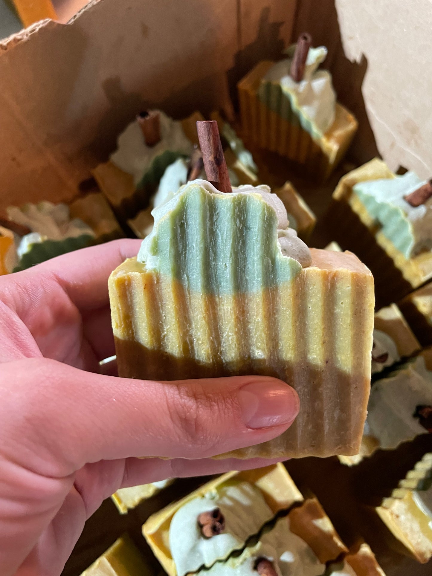 A bar of orange solid soap that is designed to look like a pumpkin. It is orange with a bit of green soap on top with a piece of cinnamon stick poking out the top. There is no label on the bar of soap. The soap is held in a hand and matching bars are present in the background. 