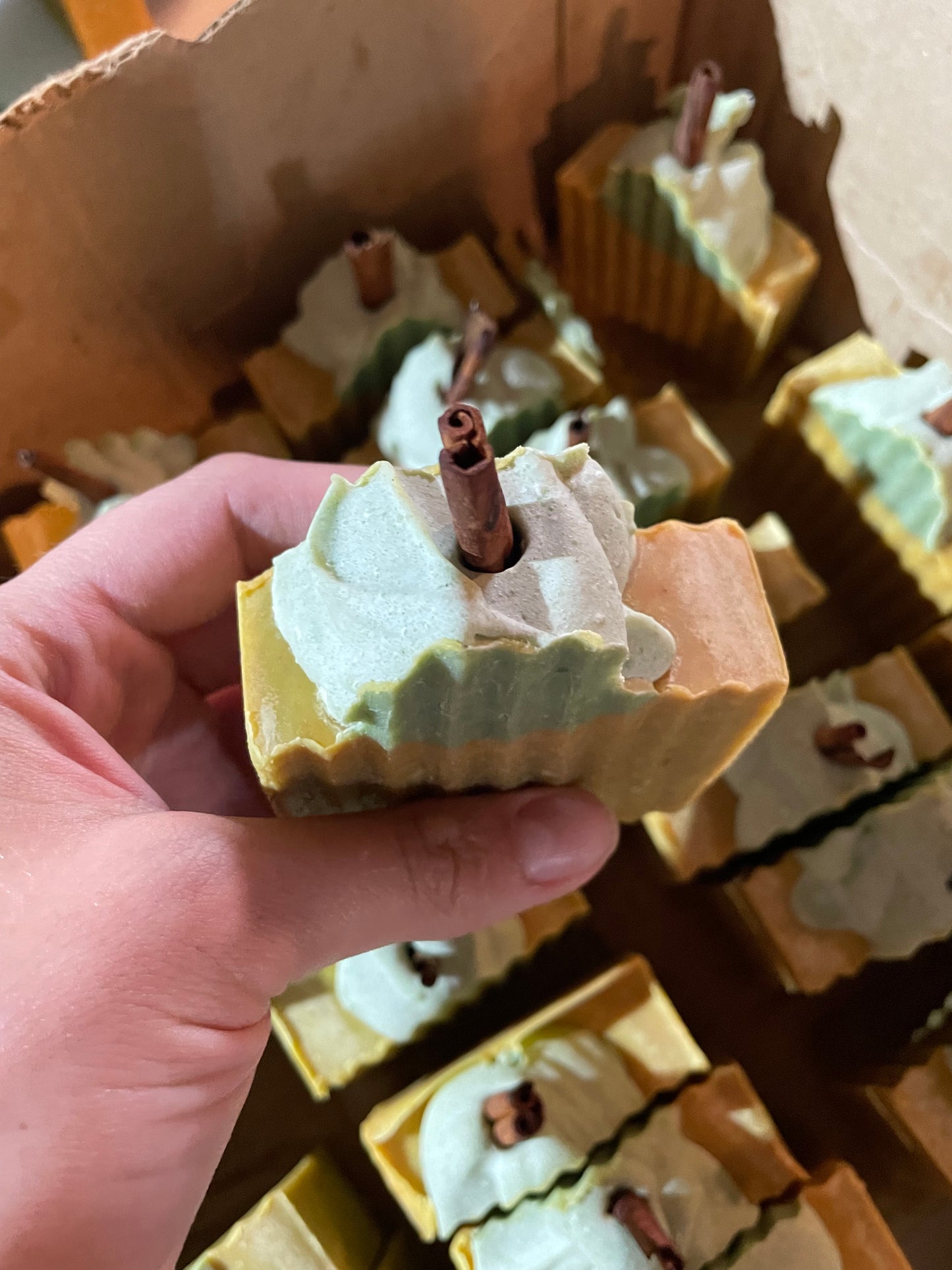 A bar of orange solid soap that is designed to look like a pumpkin. It is orange with a bit of green soap on top with a piece of cinnamon stick poking out the top. There is no label on the bar of soap. The soap is held in a hand and matching bars are present in the background. The focus of the image is the top of the soap. 