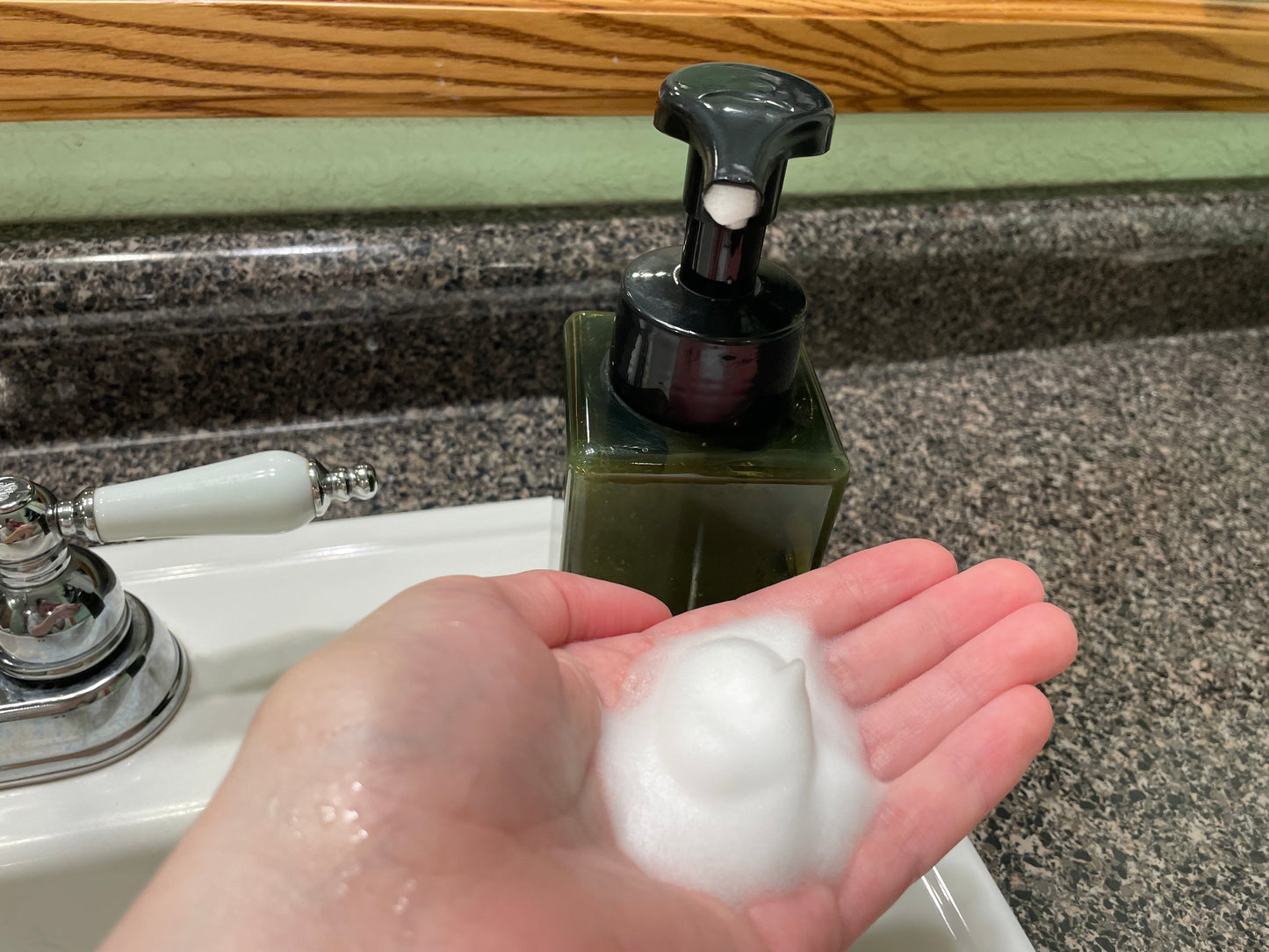 A hand with foamy liquid soap in the palm next to a black and green soap dispenser. The setting is a bathroom sink with a mirror in the background. 