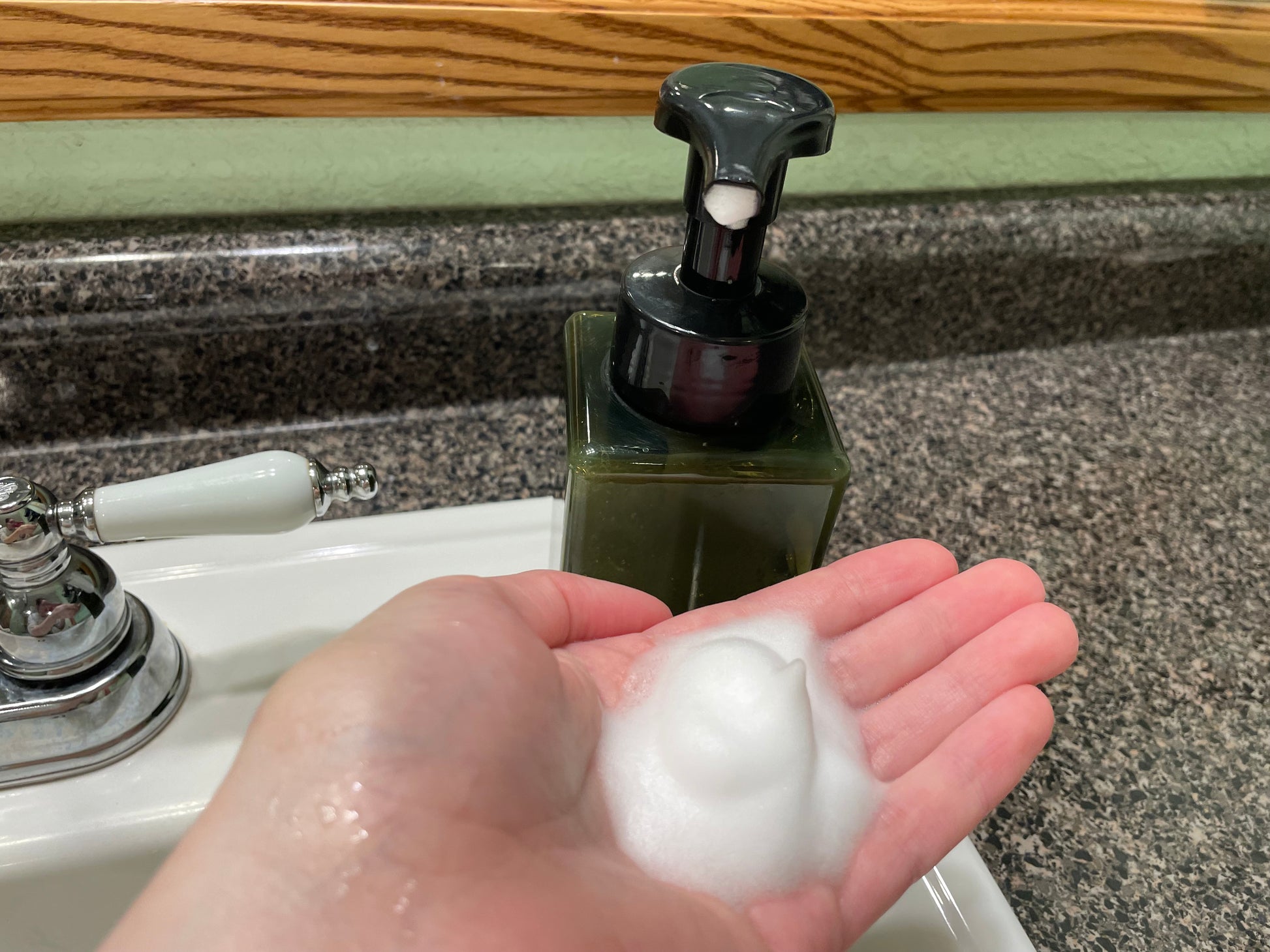 A hand with foamy liquid soap in the palm next to a black and green soap dispenser. The setting is a bathroom sink with a mirror in the background. 