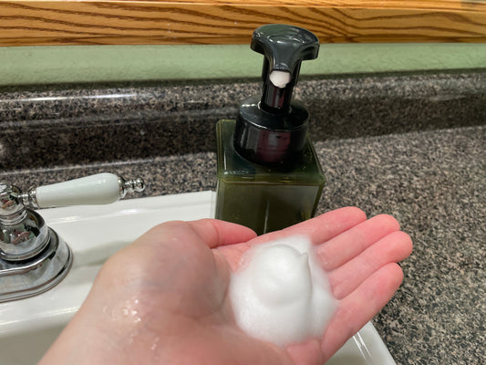 A hand with foamy liquid soap in the palm next to a black and green soap dispenser. The setting is a bathroom sink with a mirror in the background. 