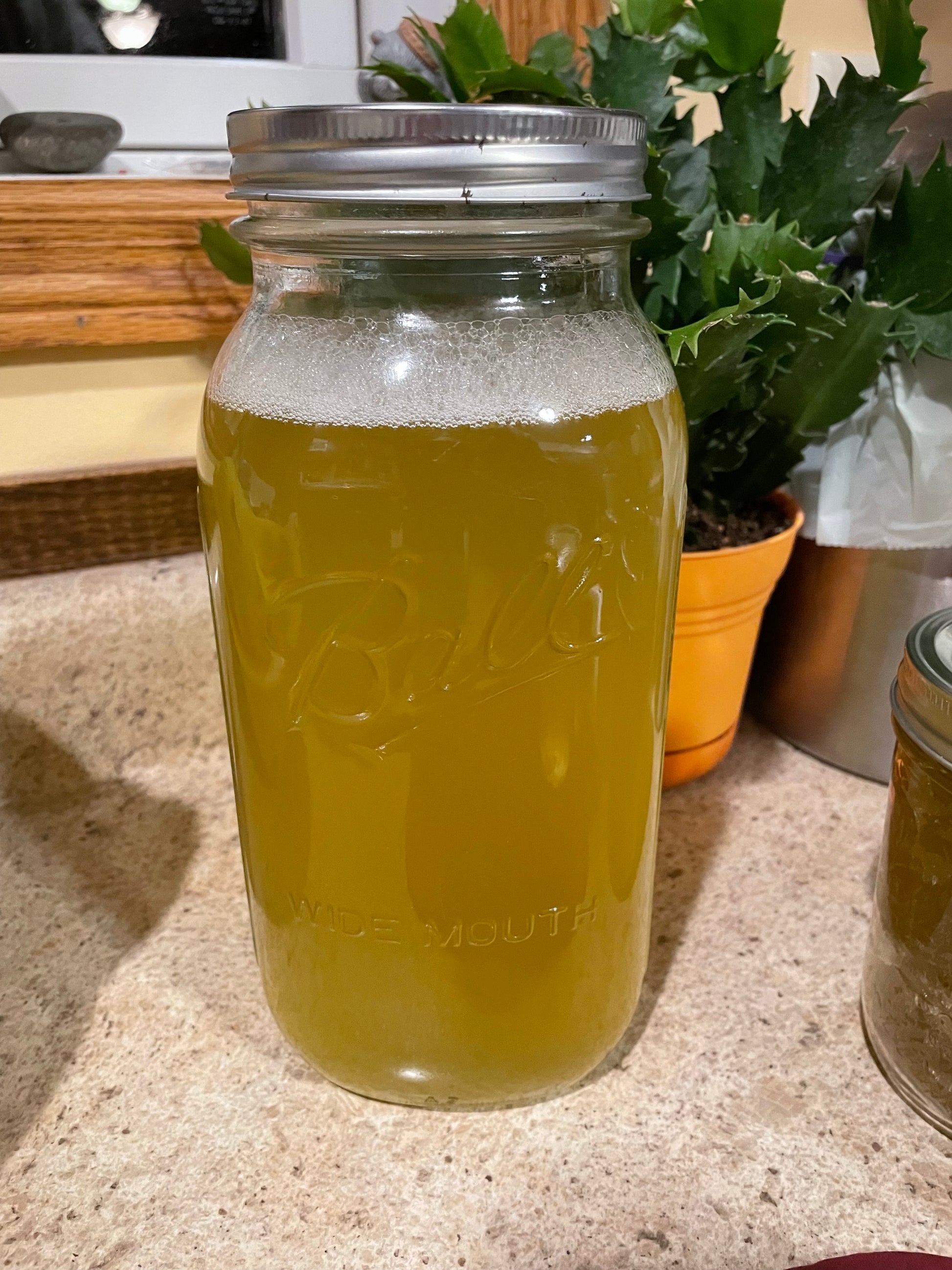 A large half a gallon jar of liquid soap concentrate. The liquid is a dark yellow color. The jar is sitting on a cream countertop. 