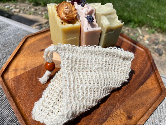 Three bars of soap with decorative elements on a wooden tray, accompanied by a mesh soap bag.