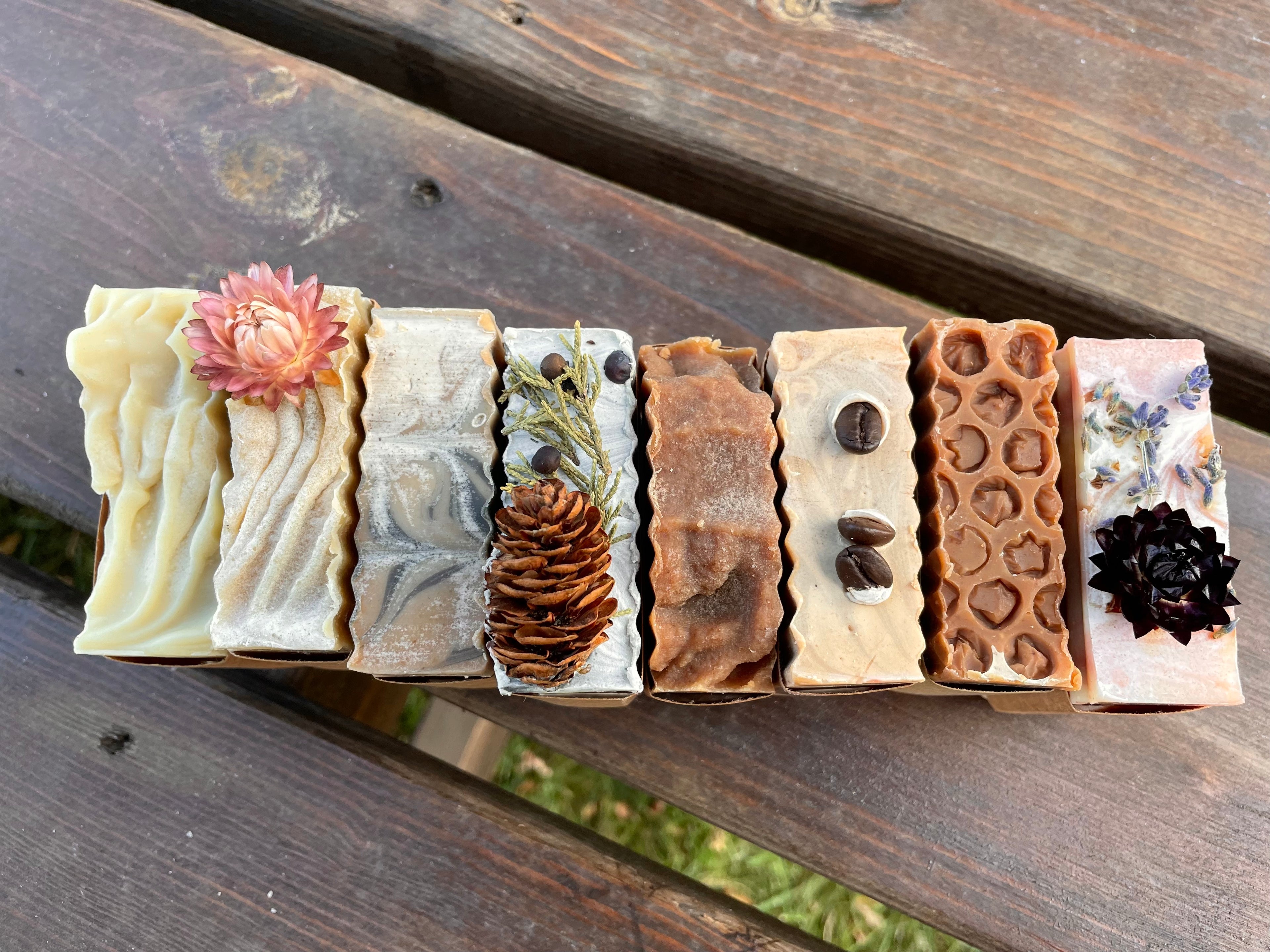 A line of hard soap bars photographed from above. Eight bars are lined up next to each other on a wood background. The soap tops range in color from white to brown. Some soaps have decorative tops including real dried flowers, coffee beans and dried pine cones. 