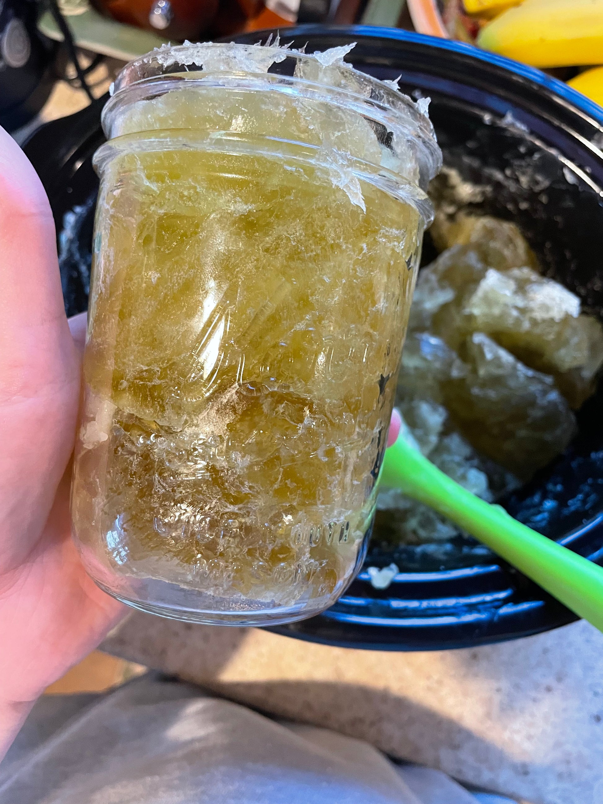 A clear glass container filled with a yellowish liquid soap concentrate, held by a hand with a blurred background.